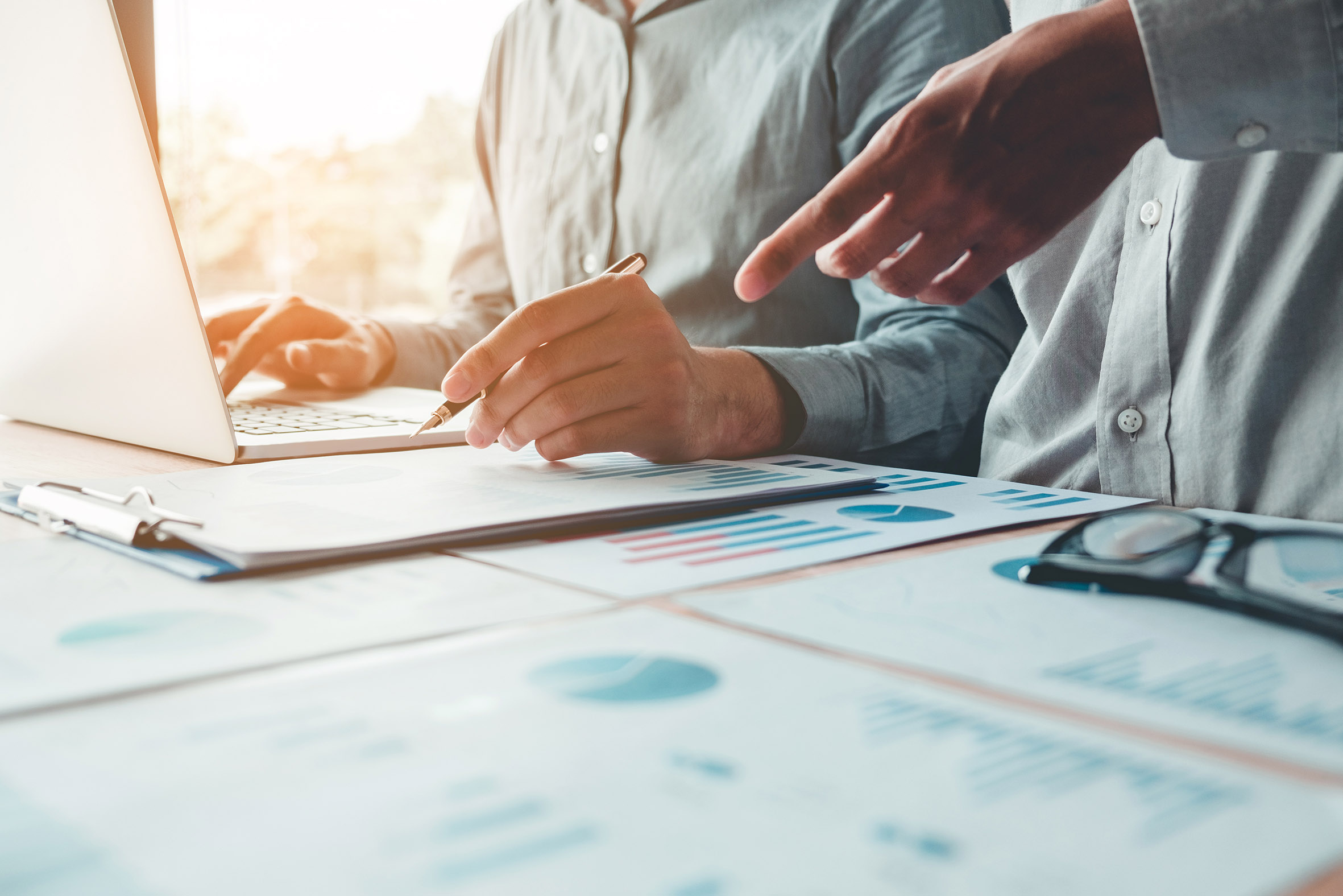 Zwei Personen arbeiten gemeinsam an einem Laptop und besprechen Auswertungen und Tabellen auf Papier.